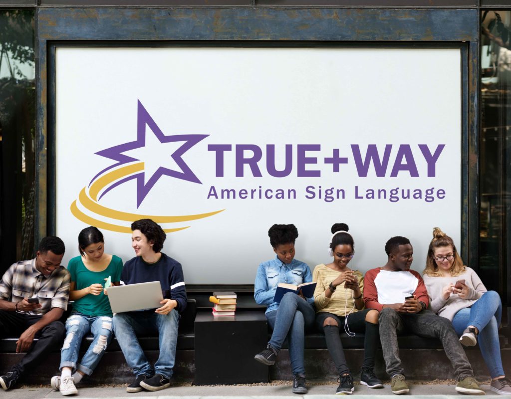 Seven diverse students sit on a bench with a large banner behind them with the TWA logo. Each student is chatting, using their phone or laptop.